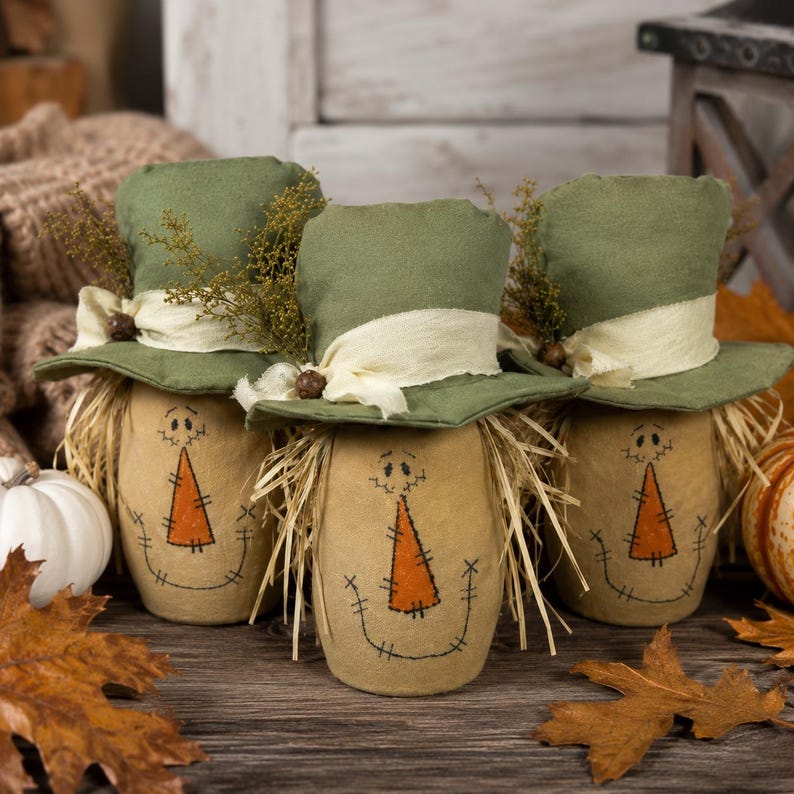 May include: Three fabric scarecrow figures with tan bodies, orange triangular noses, and stitched facial features. Each figure wears a green top hat adorned with a cream-colored fabric band, a small brown bead, and sprigs of dried greenery.