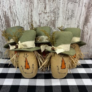 May include: A group of five fabric scarecrow figures, each featuring a tan body with a hand-drawn orange triangular nose and stitched mouth. Each figure wears a green fabric top hat adorned with a cream-colored ribbon, a small brown bell, and dried yellow foliage.