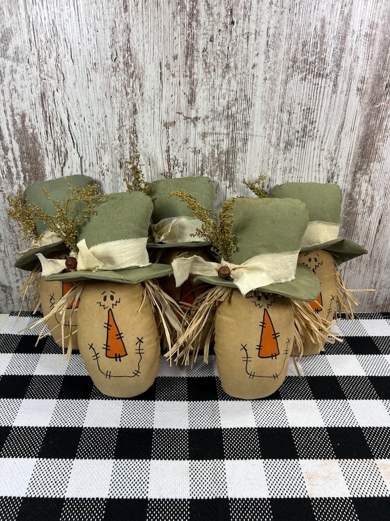 May include: A group of five fabric scarecrow figures, each featuring a tan body with a hand-drawn orange triangular nose and stitched mouth. Each figure wears a green fabric top hat adorned with a cream-colored ribbon, a small brown bell, and dried yellow foliage.