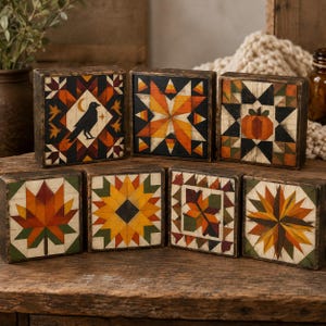 May include: A collection of seven square wooden blocks featuring painted quilt-inspired patterns in autumn colors. Designs include a black crow with a crescent moon, a star, a pumpkin, a maple leaf, a sunflower, and geometric starburst motifs.