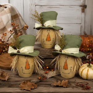 May include: Three fabric scarecrow figures with tan bodies, stitched facial features, and orange triangular noses. Each figure wears a green top hat adorned with a cream-colored ribbon, a small brown bead, and a sprig of dried foliage, with straw hair.