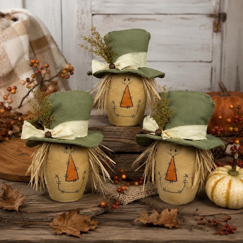 May include: Three fabric scarecrow figures with tan bodies, stitched facial features, and orange triangular noses. Each figure wears a green top hat adorned with a cream-colored ribbon, a small brown bead, and a sprig of dried foliage, with straw hair.