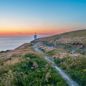 Puede incluir: Un paisaje costero escénico al anochecer. Un faro blanco con un faro iluminado se alza sobre una colina cubierta de hierba con vistas al océano. Un sendero serpentea a través de las flores silvestres verdes y moradas, bajo un cielo degradado de naranja, rosa y azul.