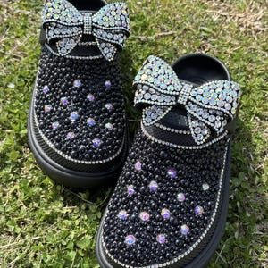 Op de afbeelding: Een paar zwarte klompen met een grote regenboogkleurige strass strik op elke schoen. De klompen zijn bedekt met kleine zwarte strass steentjes.