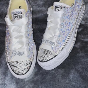 Puede incluir: Zapatillas Converse blancas con una cinta de satén blanca y un diseño de perlas blancas. Las zapatillas tienen el texto "Converse All Star" y "Grigg's". Una zapatilla tiene el texto "(1-27-99)" en ella.