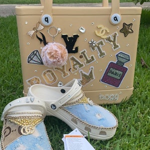 May include: A beige tote bag with a gold chain and a charm with a fluffy pom-pom. The bag is decorated with rhinestones, a crown, a star, a Chanel logo, a Louis Vuitton logo, and the word "ROYALTY". Two white clogs with blue denim and gold chain accents are in the foreground. The clogs are decorated with rhinestones and a Gucci logo.