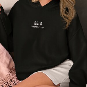 Może przedstawiać: Czarna bluza z kapturem z białym napisem "BOLD Harmony". Bluza jest w jednolitym czarnym kolorze i ma luźny krój. Obraz przedstawia tekst i fakturę materiału.