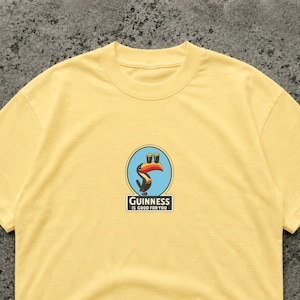 May include: A light yellow t-shirt featuring a Guinness logo. The logo includes a toucan with two pints of Guinness and the text "Guinness is good for you" in a rectangular box. The shirt is made of a soft material.
