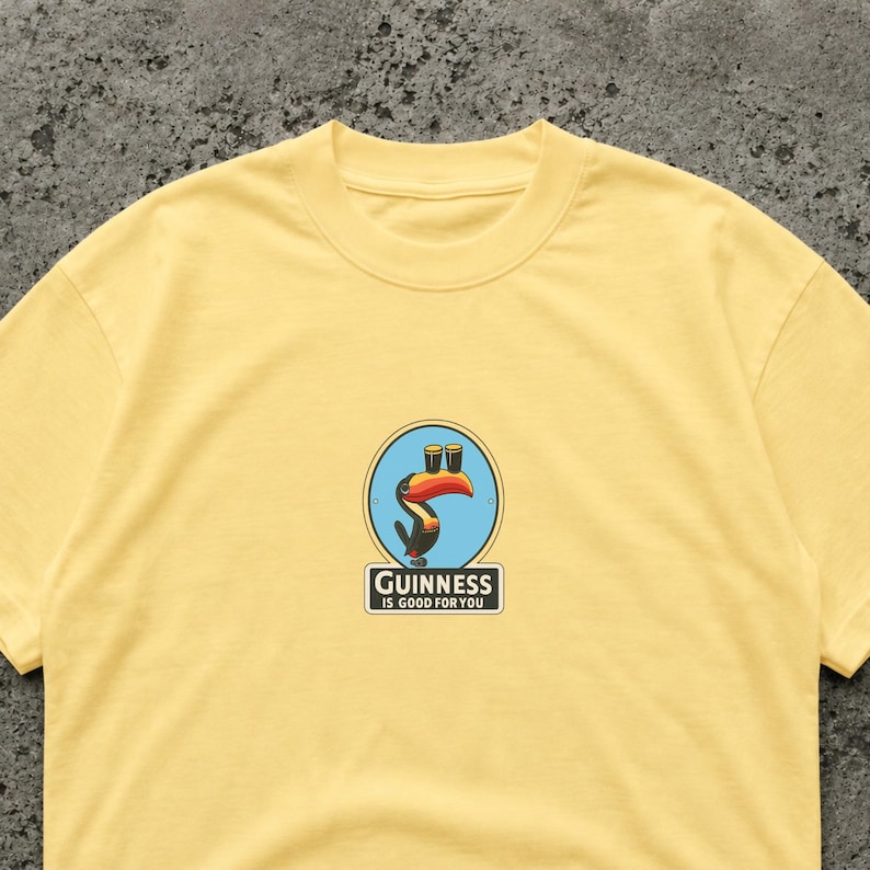 May include: A light yellow t-shirt featuring a Guinness logo. The logo includes a toucan with two pints of Guinness and the text "Guinness is good for you" in a rectangular box. The shirt is made of a soft material.