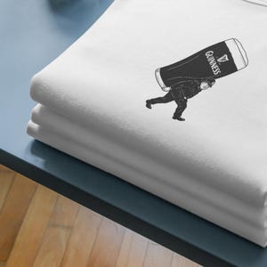 Puede incluir: Camisetas blancas dobladas con un gr&aacute;fico negro de una persona que lleva una pinta grande de Guinness. El logotipo de Guinness es visible en la pinta. Las camisetas est&aacute;n apiladas sobre una superficie azul.