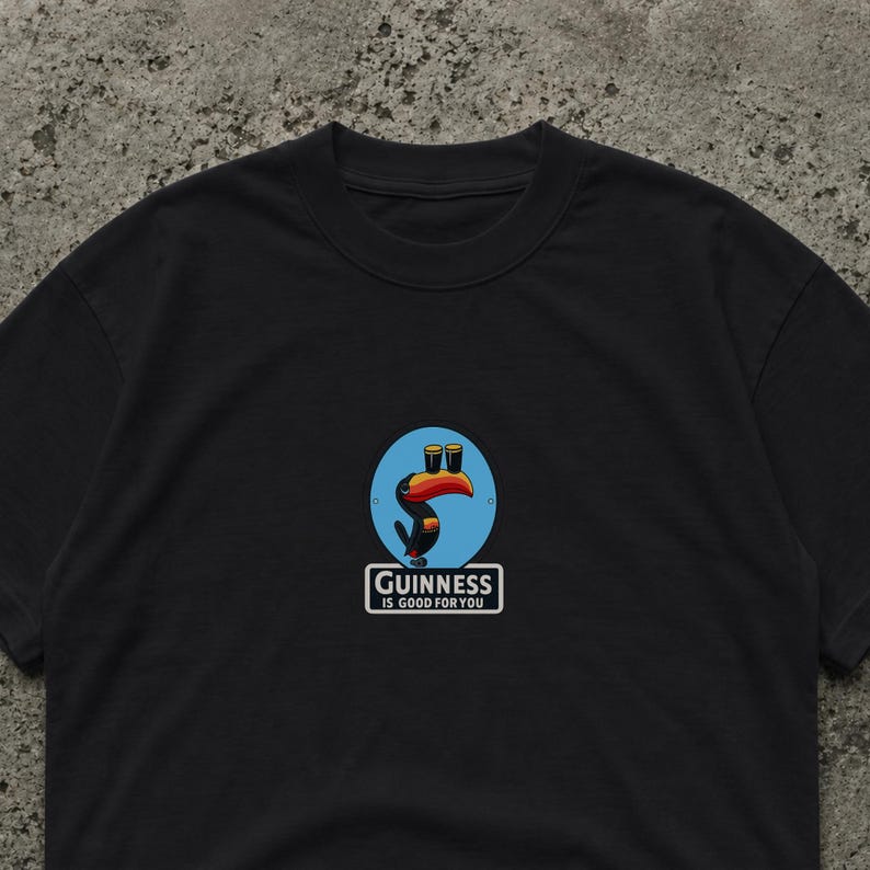 Op de afbeelding: Zwart t-shirt met een cartoon toekan die twee pinten bier op zijn snavel draagt, tegen een blauwe cirkel. De tekst "Guinness is good for you" staat in een witte rechthoek.