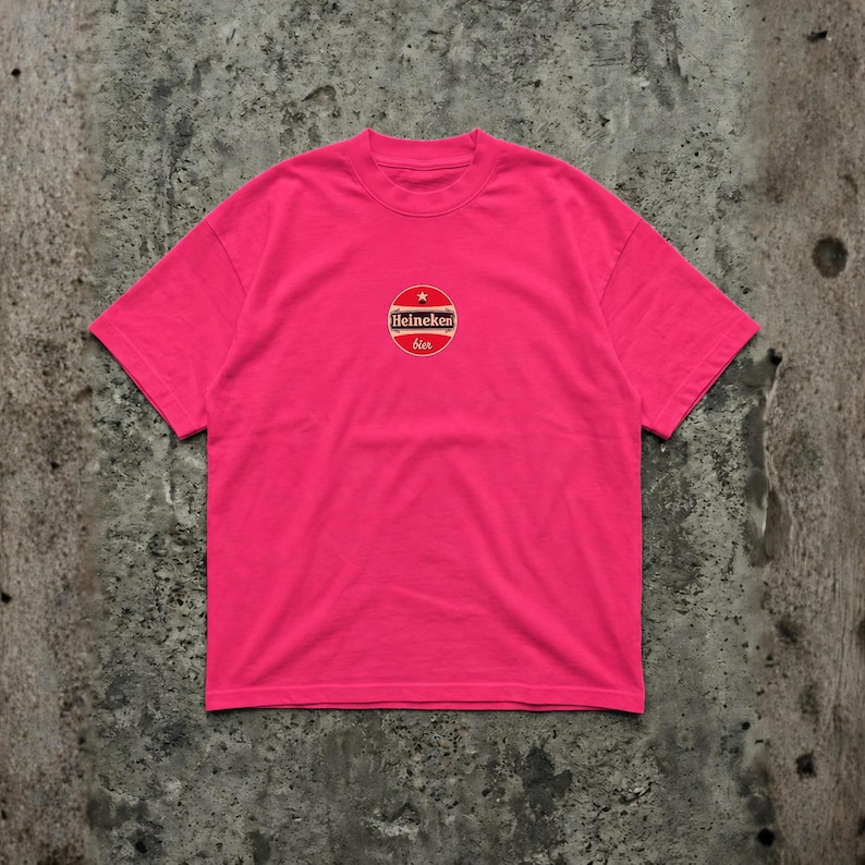 Pu&ograve; includere: Una maglietta rosa acceso con girocollo e maniche corte. Presenta un logo circolare con una stella rossa e le parole "Heineken" e "Bier" in oro. La maglietta &egrave; realizzata in tessuto morbido.