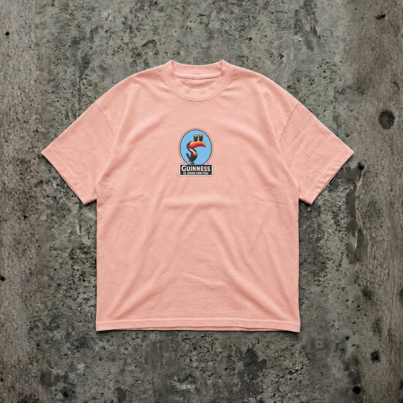 K&ouml;nnte beinhalten: Hellrosa T-Shirt mit einem Guinness-Logo, das einen Tukan in einem blauen Kreis zeigt. Das Logo enth&auml;lt den Text "Guinness is good for you". Die Grafik ist blau, orange und schwarz.