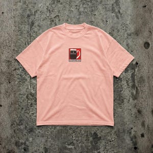 May include: A light pink t-shirt featuring a small Guinness logo on the front. The logo is a stylized pint of Guinness with the brand name below. The shirt has a crew neck and short sleeves.