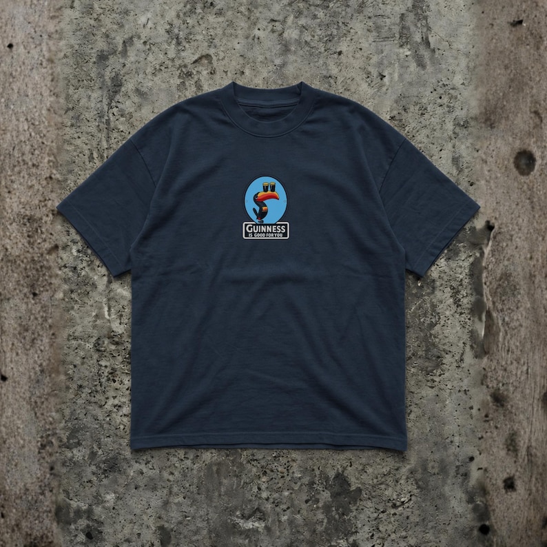 K&ouml;nnte beinhalten: Marineblaues T-Shirt mit einem kreisf&ouml;rmigen Logo, das einen Tukan, zwei blaue Gl&auml;ser und den Text "Guinness" zeigt. Das Logo ist mittig auf der Brust platziert. Das Shirt hat kurze &Auml;rmel und einen Rundhalsausschnitt.