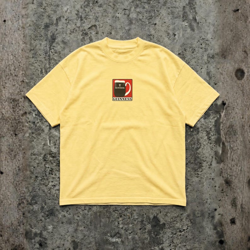 May include: Yellow t-shirt featuring a Guinness logo on the front. The logo is a red and white design with the word "Guinness" below it. The shirt has short sleeves and a crew neck.