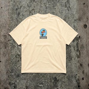 K&ouml;nnte beinhalten: Cremefarbenes T-Shirt mit einem kreisf&ouml;rmigen Aufdruck, der einen Tukan, ein Pint Bier und den Text "Guinness is good for you" zeigt. Die Grafik ist in Blau, Orange und Schwarz gehalten. Das Shirt hat kurze &Auml;rmel und einen Rundhalsausschnitt.