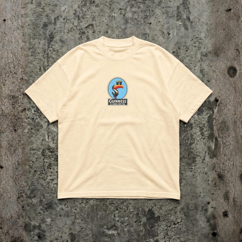K&ouml;nnte beinhalten: Cremefarbenes T-Shirt mit einem kreisf&ouml;rmigen Aufdruck, der einen Tukan, ein Pint Bier und den Text "Guinness is good for you" zeigt. Die Grafik ist in Blau, Orange und Schwarz gehalten. Das Shirt hat kurze &Auml;rmel und einen Rundhalsausschnitt.