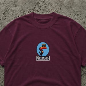 May include: Burgundy t-shirt featuring a circular graphic with a toucan holding two pints of stout. The graphic is set against a blue background. The text "Guinness is good for you" is below the graphic.