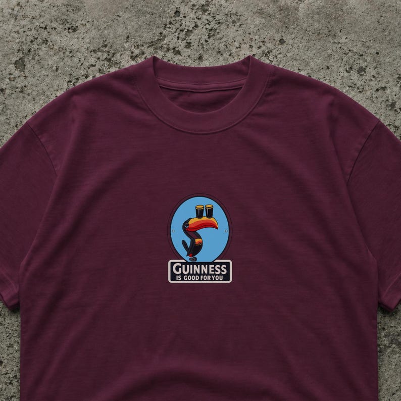 May include: Burgundy t-shirt featuring a circular graphic with a toucan holding two pints of stout. The graphic is set against a blue background. The text "Guinness is good for you" is below the graphic.