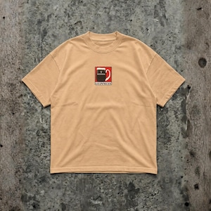 May include: A tan-colored t-shirt featuring a small graphic of a Guinness pint glass. The graphic is in red, black, and white, with the Guinness logo below. The shirt has a crew neck and short sleeves.