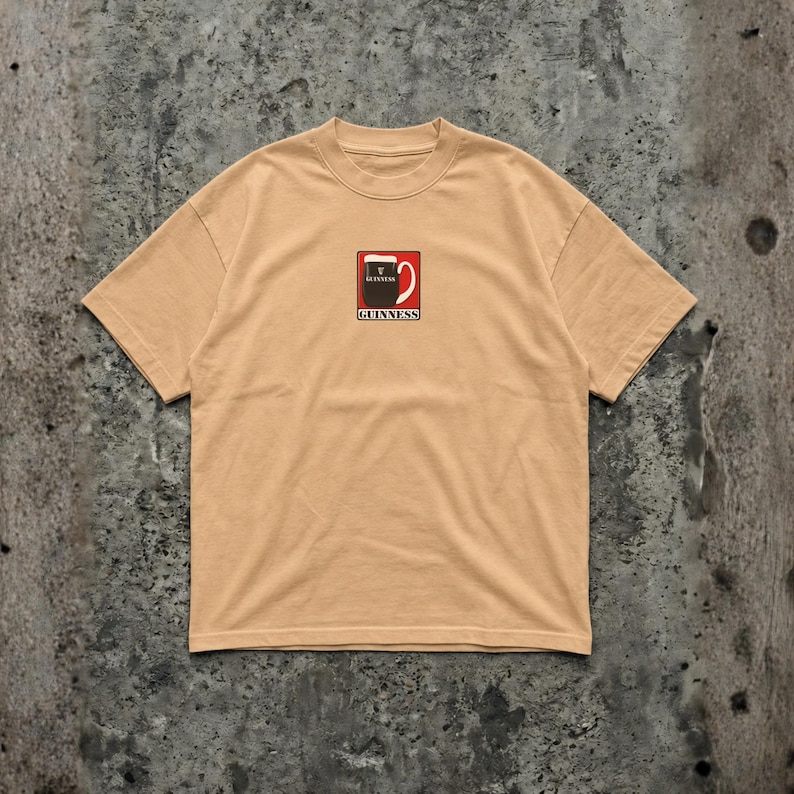 May include: A tan-colored t-shirt featuring a small graphic of a Guinness pint glass. The graphic is in red, black, and white, with the Guinness logo below. The shirt has a crew neck and short sleeves.