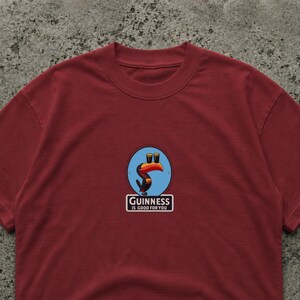 May include: Burgundy crew neck t-shirt with a circular graphic featuring a toucan with two pints of beer and the text "Guinness is good for you". The graphic is in blue, orange, and black.