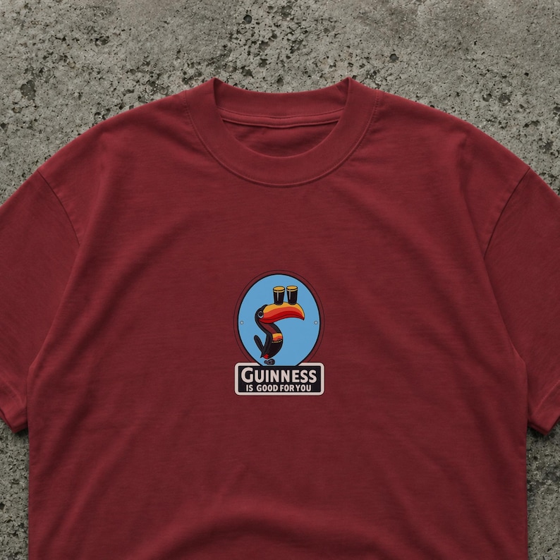 May include: Burgundy crew neck t-shirt with a circular graphic featuring a toucan with two pints of beer and the text "Guinness is good for you". The graphic is in blue, orange, and black.