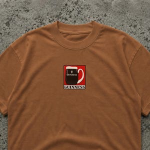May include: A brown t-shirt with a Guinness pint graphic in a red square. The Guinness logo is above and below the pint illustration. The shirt has a crew neck and is made of a soft material.