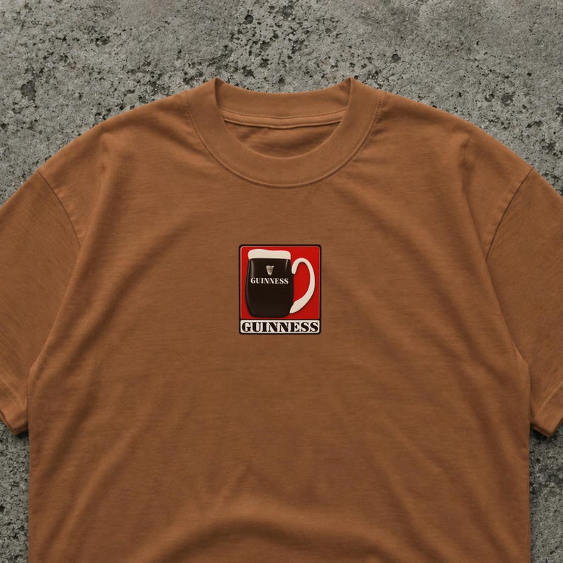May include: A brown t-shirt with a Guinness pint graphic in a red square. The Guinness logo is above and below the pint illustration. The shirt has a crew neck and is made of a soft material.