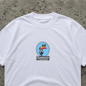 May include: White t-shirt featuring a colorful Guinness logo with a toucan and two pints of beer. The text "Guinness is good for you" is printed below the logo. The shirt is made of a soft material.