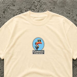 May include: Cream-colored t-shirt featuring a circular graphic with a toucan holding two pints of beer. The graphic is surrounded by a blue border and the text "Guinness is good for you" is below the graphic.