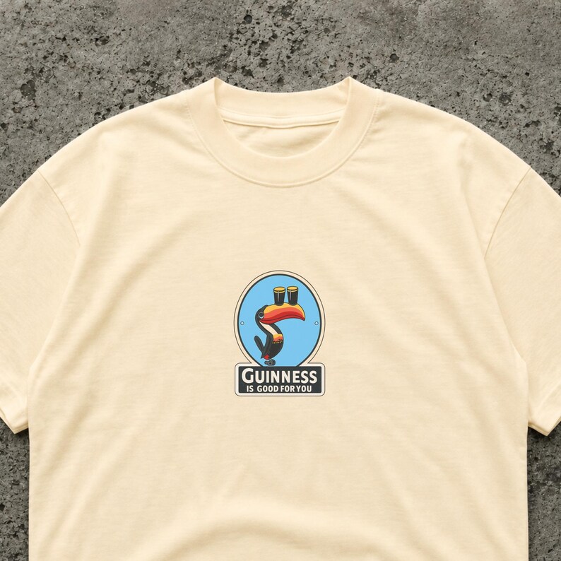 May include: Cream-colored t-shirt featuring a circular graphic with a toucan holding two pints of beer. The graphic is surrounded by a blue border and the text "Guinness is good for you" is below the graphic.
