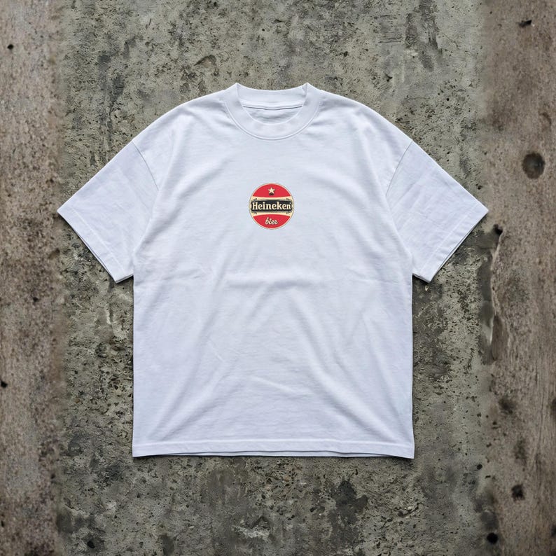 Pu&ograve; includere: T-shirt bianco con girocollo e maniche corte. La maglietta presenta un logo circolare al centro con colori rosso, bianco e oro. Il logo include il testo "Heineken" e "bier".