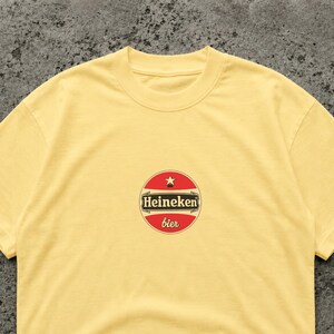 Puede incluir: Una camiseta amarilla clara con un logotipo circular. El logotipo muestra el texto "Heineken bier" en rojo y blanco. La camiseta tiene cuello redondo y mangas cortas, adecuada para ropa casual.