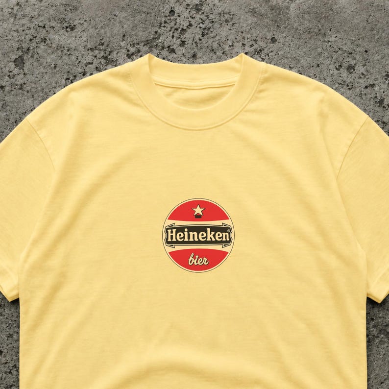 Puede incluir: Una camiseta amarilla clara con un logotipo circular. El logotipo muestra el texto "Heineken bier" en rojo y blanco. La camiseta tiene cuello redondo y mangas cortas, adecuada para ropa casual.