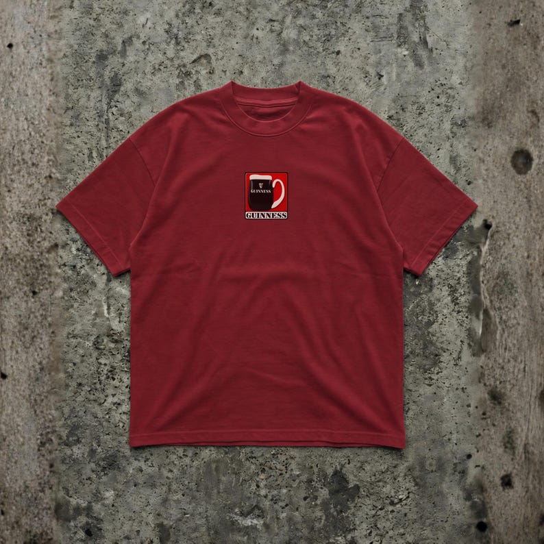 May include: A maroon t-shirt featuring a small graphic of a Guinness pint glass with the brand name below. The shirt has short sleeves and a crew neck. The graphic is in red, white, and black.