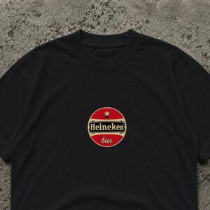 Peut inclure: T-shirt noir avec un logo circulaire "Heineken bier". Le logo est rouge avec un bord dor&eacute; et une &eacute;toile. Le t-shirt est en mati&egrave;re souple. Le texte "bier" est en &eacute;criture cursive.