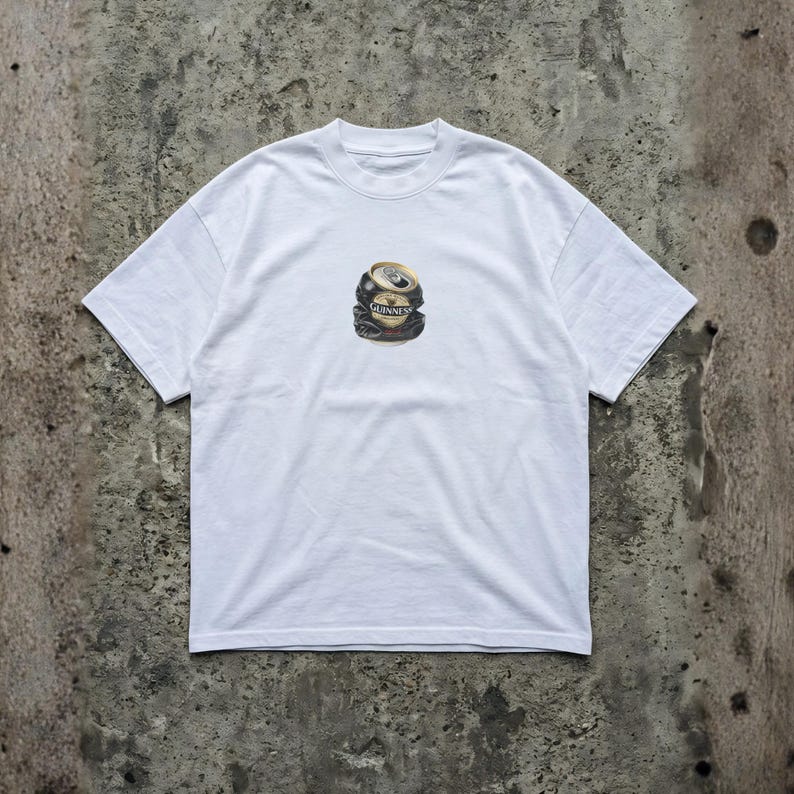 May include: White t-shirt featuring a graphic of a crushed Guinness beer can. The can is black, gold, and silver, with the Guinness logo visible. The shirt has short sleeves and a crew neck.