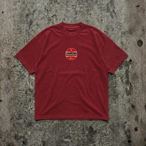 Pu&ograve; includere: Una maglietta bordeaux con girocollo e maniche corte. La maglietta presenta un logo circolare con il testo "Heineken" e "bier" in un design oro e rosso. La maglietta &egrave; realizzata in un materiale morbido e confortevole.
