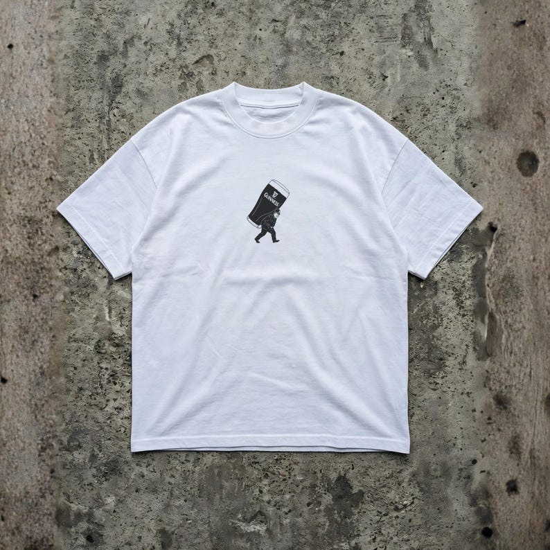 Puede incluir: Camiseta blanca con un gr&aacute;fico en blanco y negro de una persona que lleva una pinta grande de Guinness. El logotipo de Guinness es visible en la pinta. La camiseta tiene mangas cortas y cuello redondo.