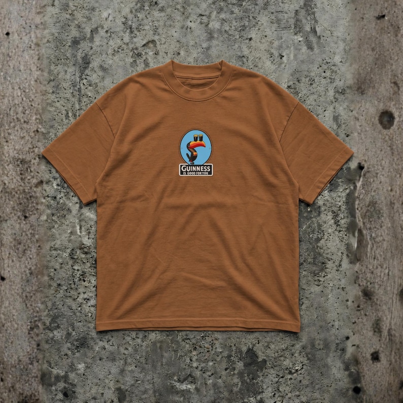 K&ouml;nnte beinhalten: Ein braunes T-Shirt mit einem kreisf&ouml;rmigen Logo, das einen Tukan, einen blauen Hintergrund und den Text "Guinness" in einem wei&szlig;en Rechteck zeigt. Das T-Shirt hat kurze &Auml;rmel und einen Rundhalsausschnitt.
