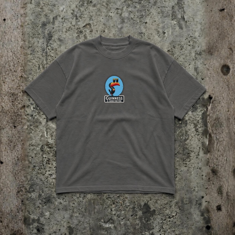 K&ouml;nnte beinhalten: Graues T-Shirt mit einem Guinness-Logo mit einem Tukan und Text. Das Logo befindet sich in einem blau-wei&szlig;en Kreis. Der Text "Guinness is good for you" steht darunter. Das Shirt ist aus weichem Material.