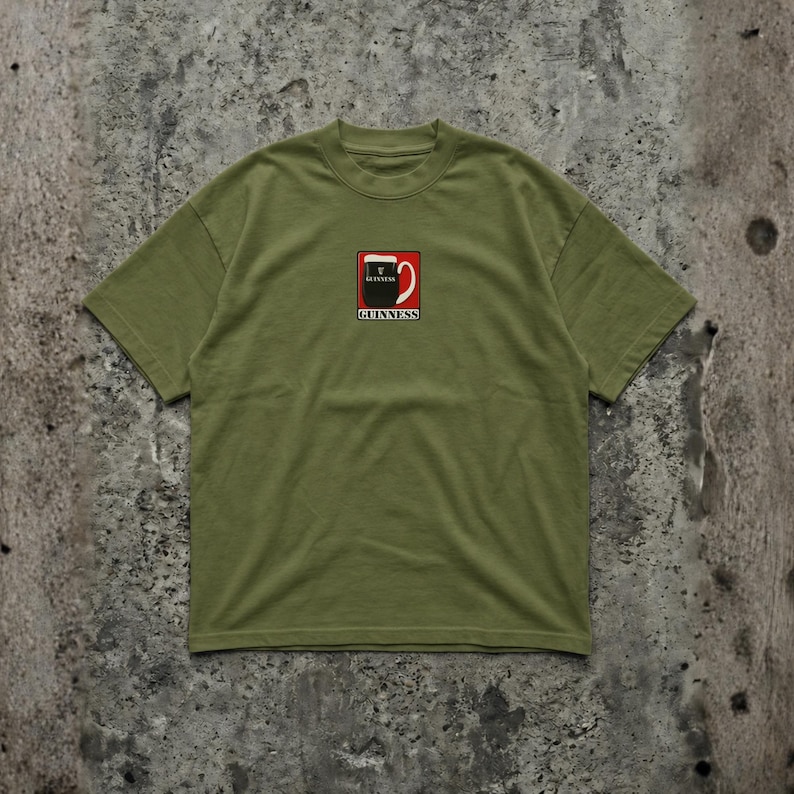 May include: Olive green t-shirt featuring a square graphic of a Guinness pint glass with the brand name below. The graphic is red, black, and white. The shirt has short sleeves and a crew neck.