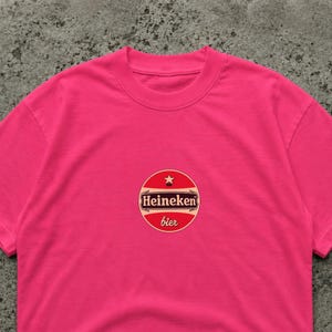K&ouml;nnte beinhalten: Ein leuchtend pinkfarbenes T-Shirt mit einem kreisf&ouml;rmigen Logo "Heineken bier". Das Logo ist rot, wei&szlig; und gold, mit einem Stern. Das T-Shirt ist einfarbig und hat einen Rundhalsausschnitt.