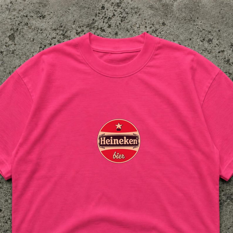 K&ouml;nnte beinhalten: Ein leuchtend pinkfarbenes T-Shirt mit einem kreisf&ouml;rmigen Logo "Heineken bier". Das Logo ist rot, wei&szlig; und gold, mit einem Stern. Das T-Shirt ist einfarbig und hat einen Rundhalsausschnitt.