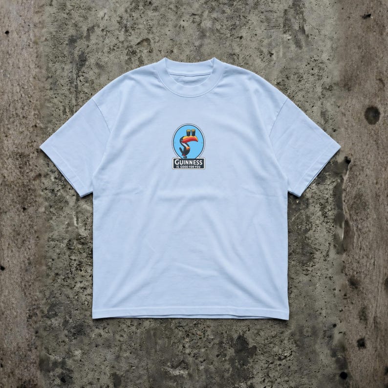 K&ouml;nnte beinhalten: Hellblaues T-Shirt mit einem kreisf&ouml;rmigen Grafikdesign, das einen Tukan und das Guinness-Logo zeigt. Die Grafik ist in Rot-, Orange- und Blaut&ouml;nen gehalten. Das Shirt besteht aus weichem Material und hat einen Rundhalsausschnitt.