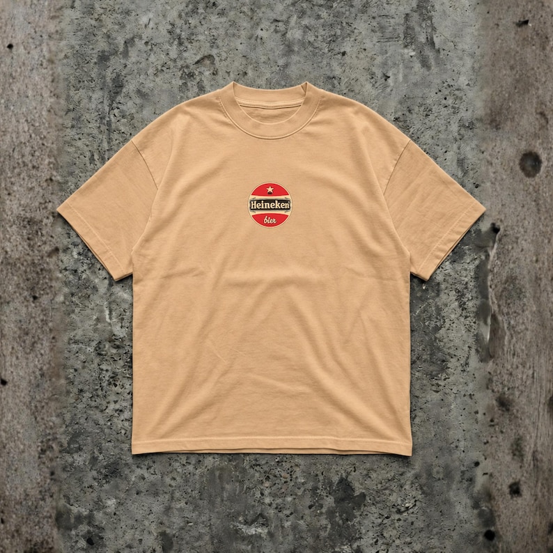 Pu&ograve; includere: Una maglietta beige con un logo circolare con la scritta "Heineken bier". Il logo &egrave; rosso, bianco e verde, con una stella in alto. La maglietta ha un girocollo e maniche corte.