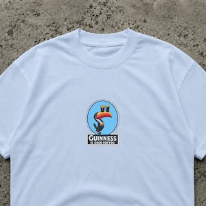 Puede incluir: Camiseta blanca con un dise&ntilde;o circular que muestra un tuc&aacute;n sosteniendo dos pintas de Guinness. El dise&ntilde;o est&aacute; sobre un fondo azul, con el texto "Guinness is good for you" debajo.