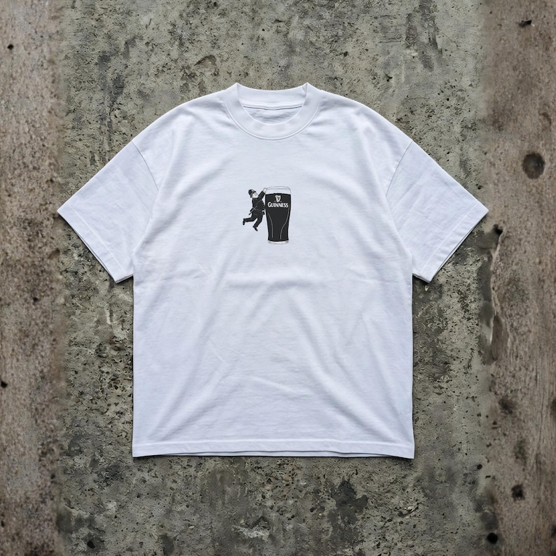 May include: White t-shirt featuring a black graphic of a person climbing a pint of Guinness. The Guinness logo is visible on the pint. The shirt has short sleeves and a crew neck.